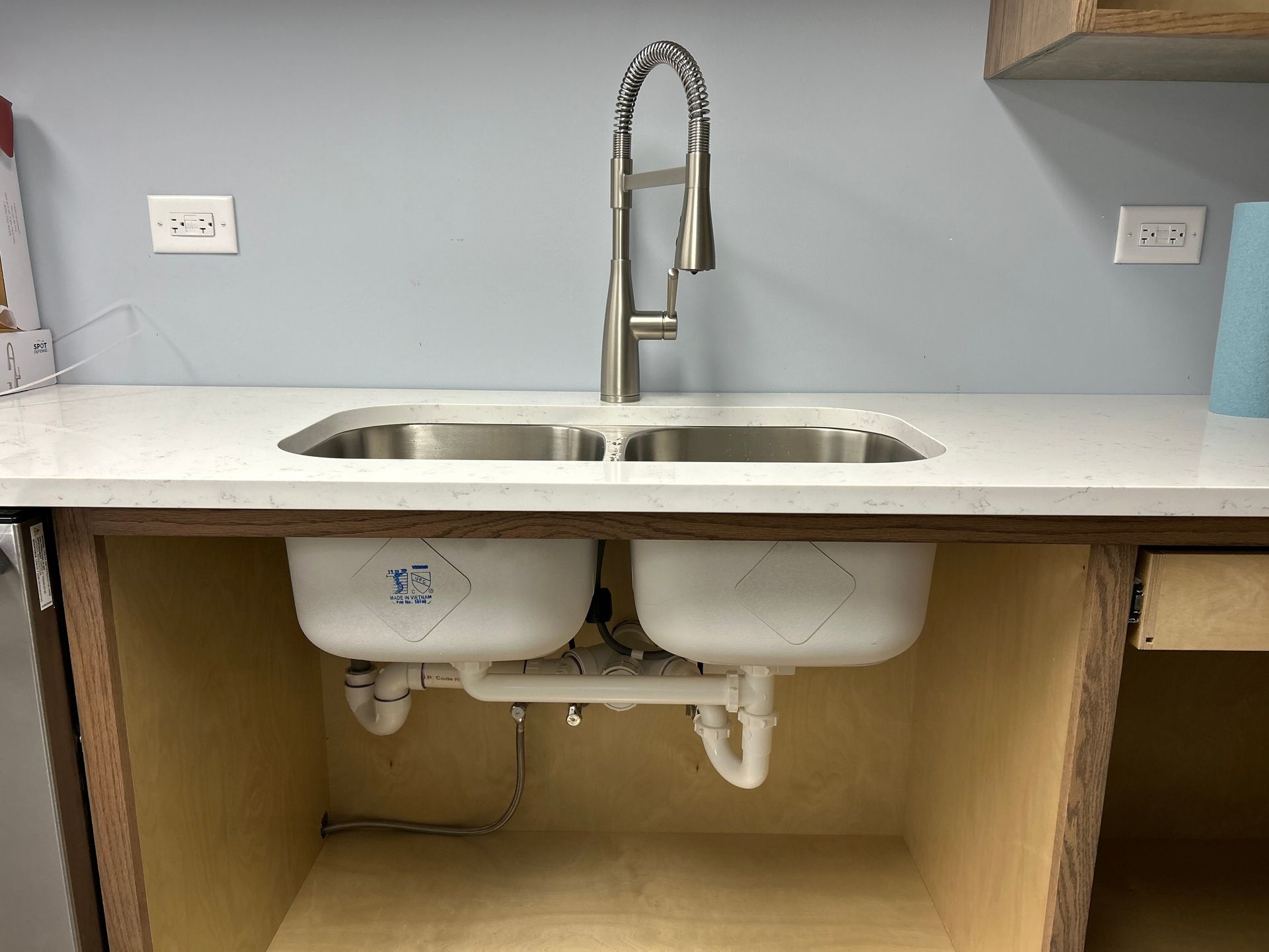Kitchen sink with two basins and a tall faucet, mounted in a light-colored countertop, with exposed cabinetry below.