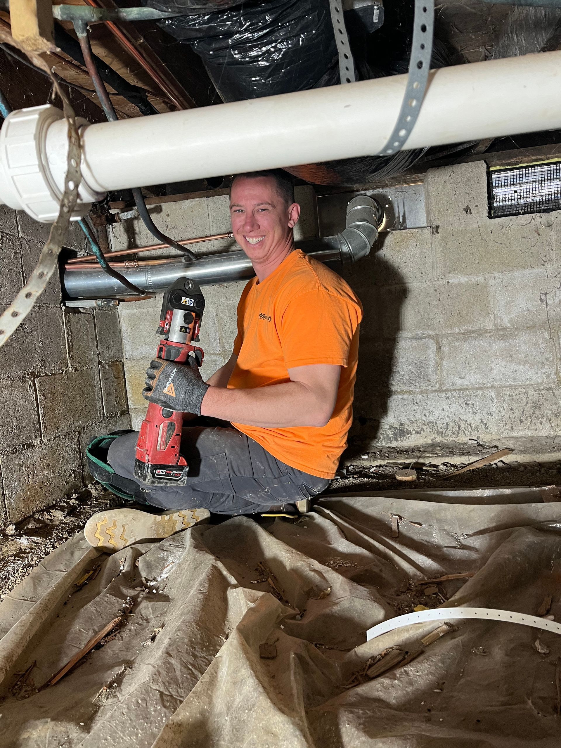 Man in orange shirt kneels under pipes, using a drill. Dark, damp crawlspace setting.