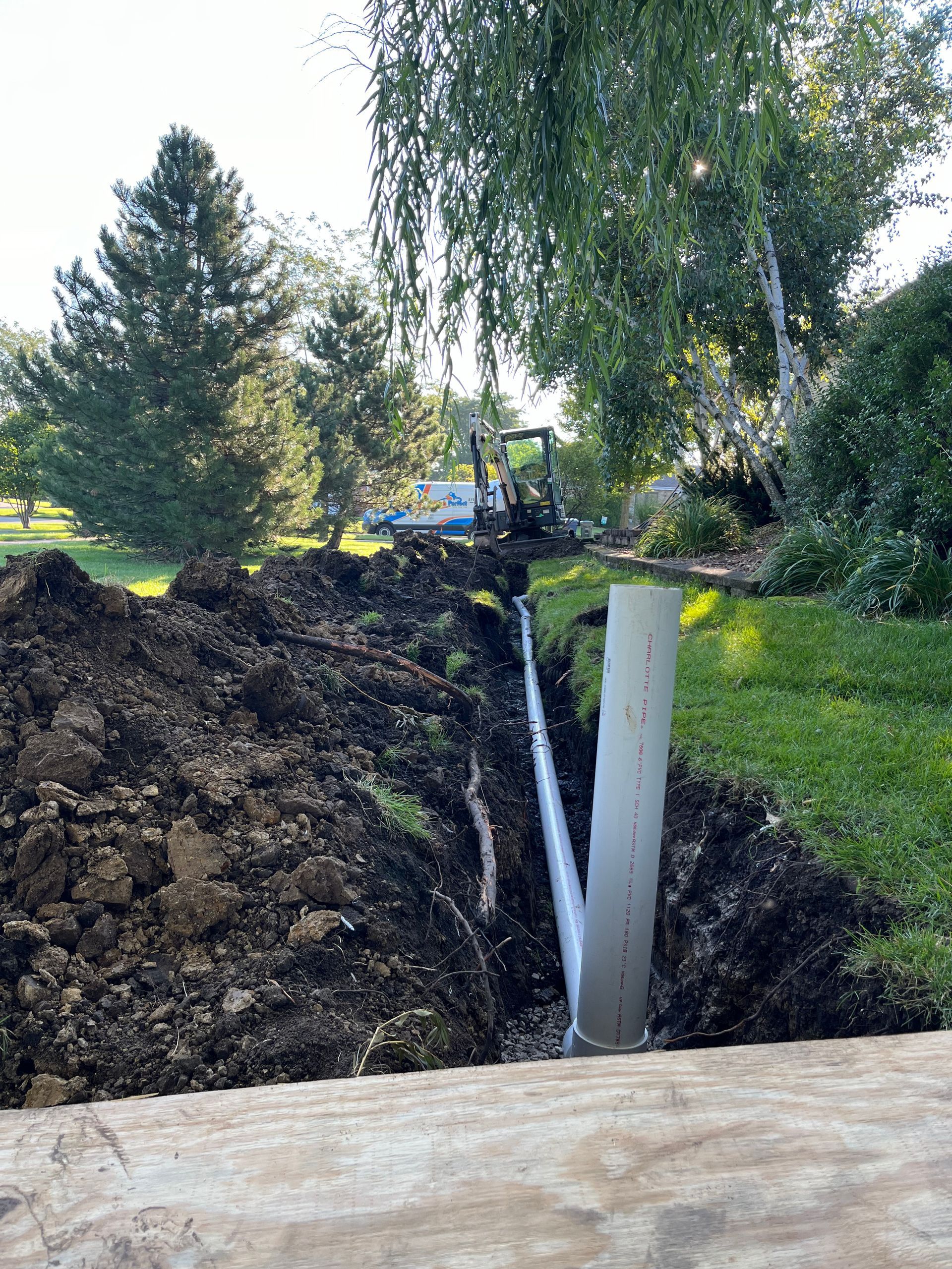 Trench dug in yard with a pipe shooting water; excavator in the background.