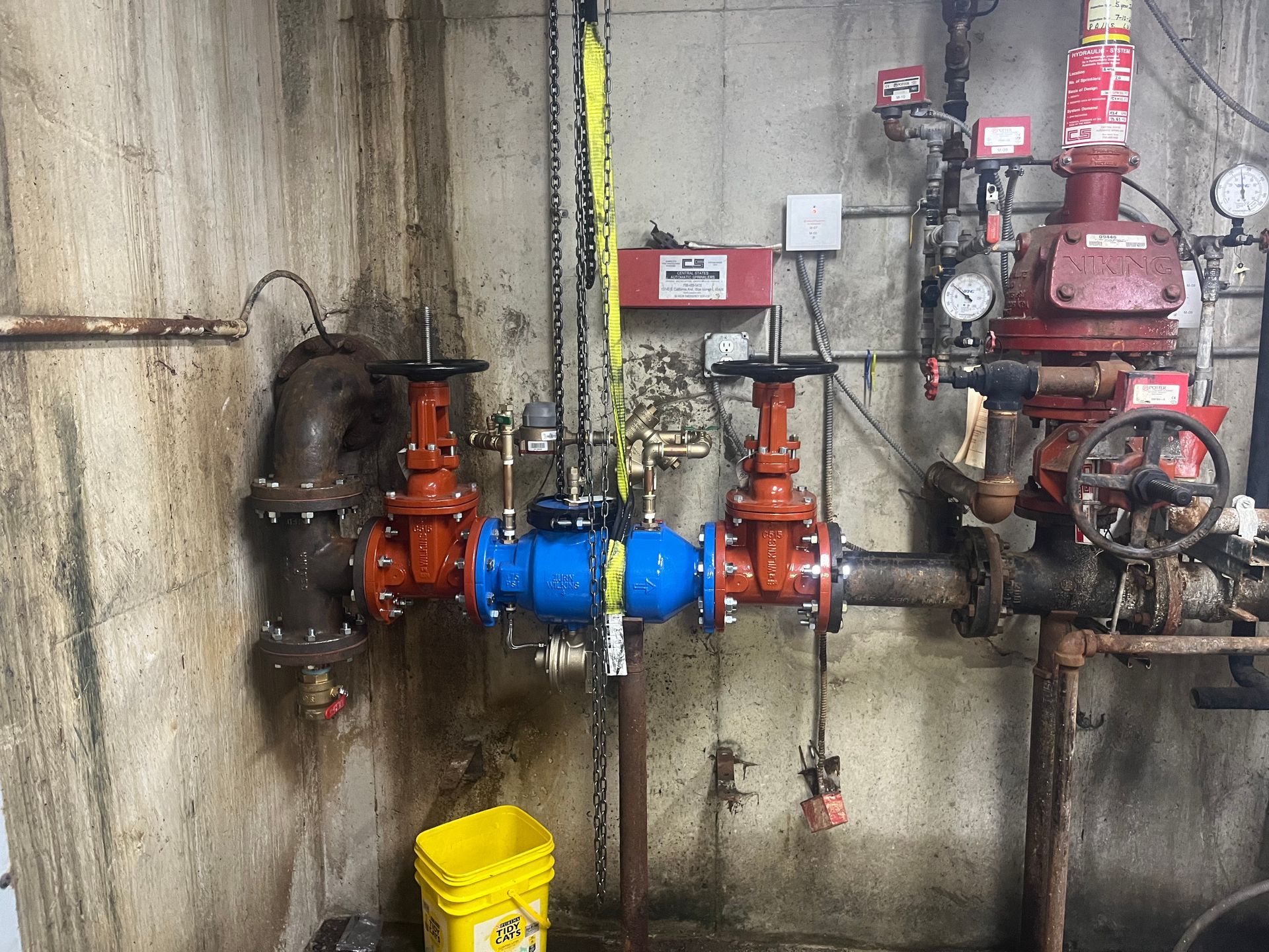 Plumbing system with red and blue valves, pipes, and gauges in a concrete basement.