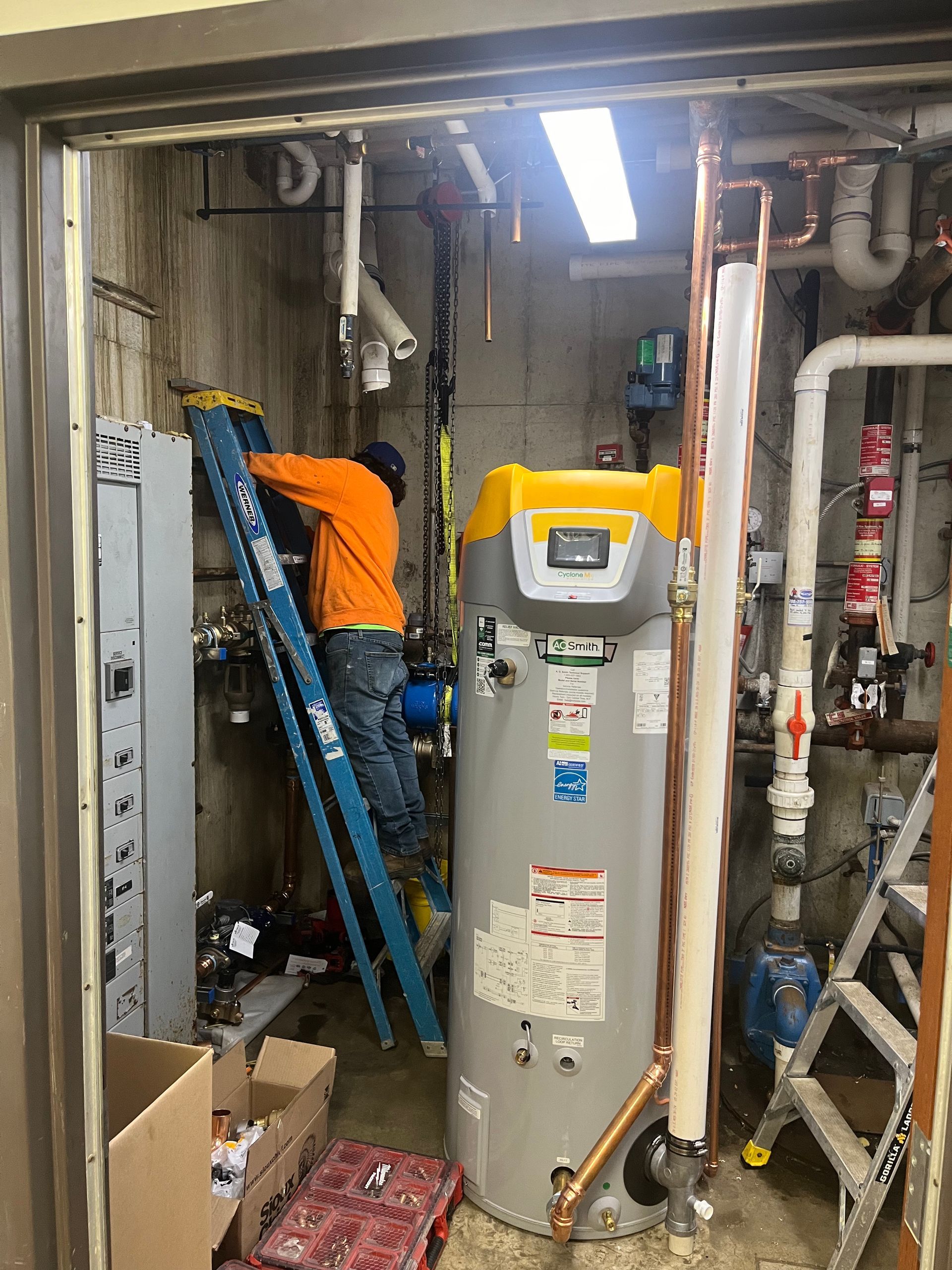 A man is standing on a ladder in a room next to a water heater.
