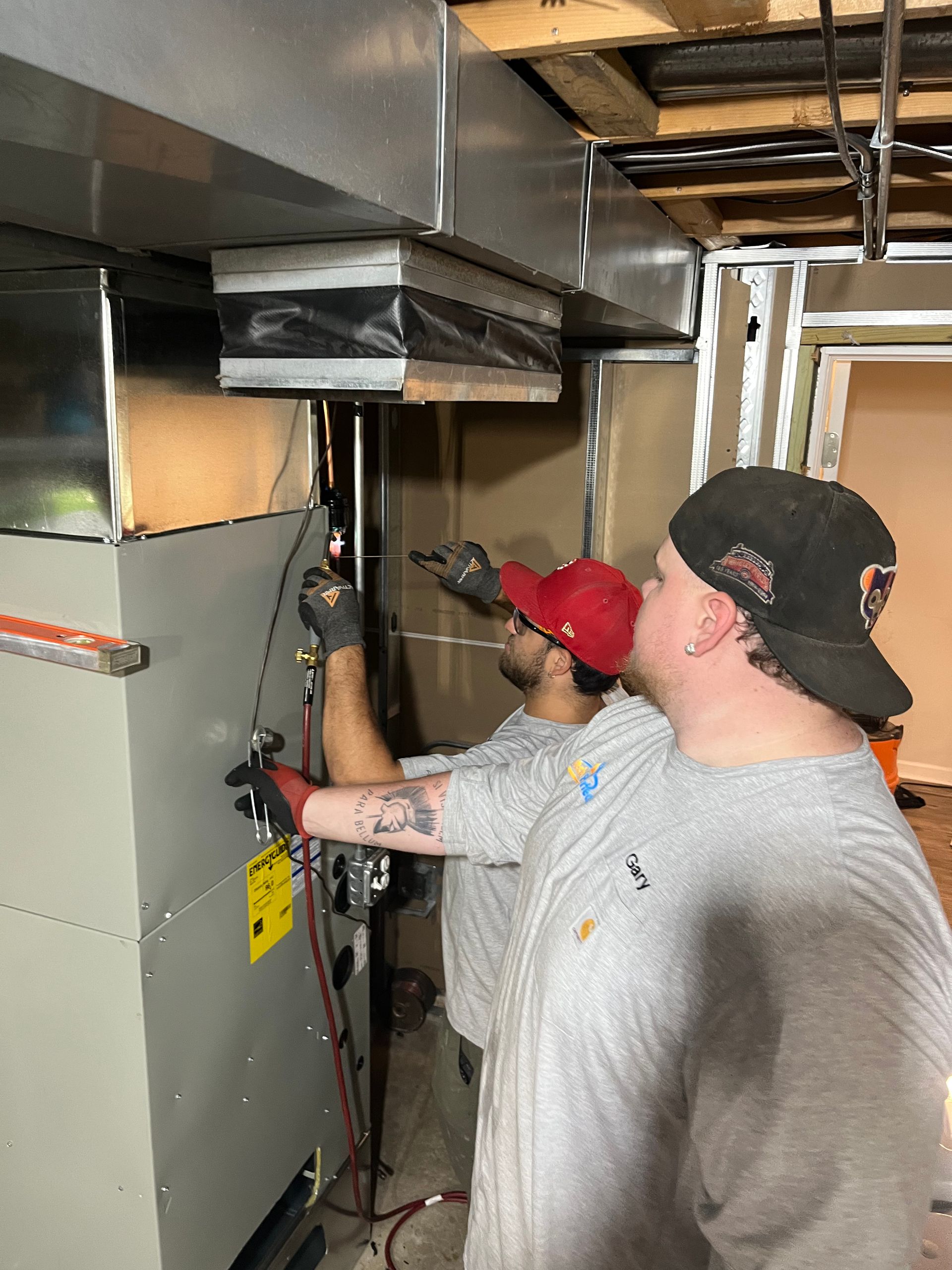 Two workers installing HVAC system in a basement. One in a red cap, the other in a black cap, both wearing gray shirts.