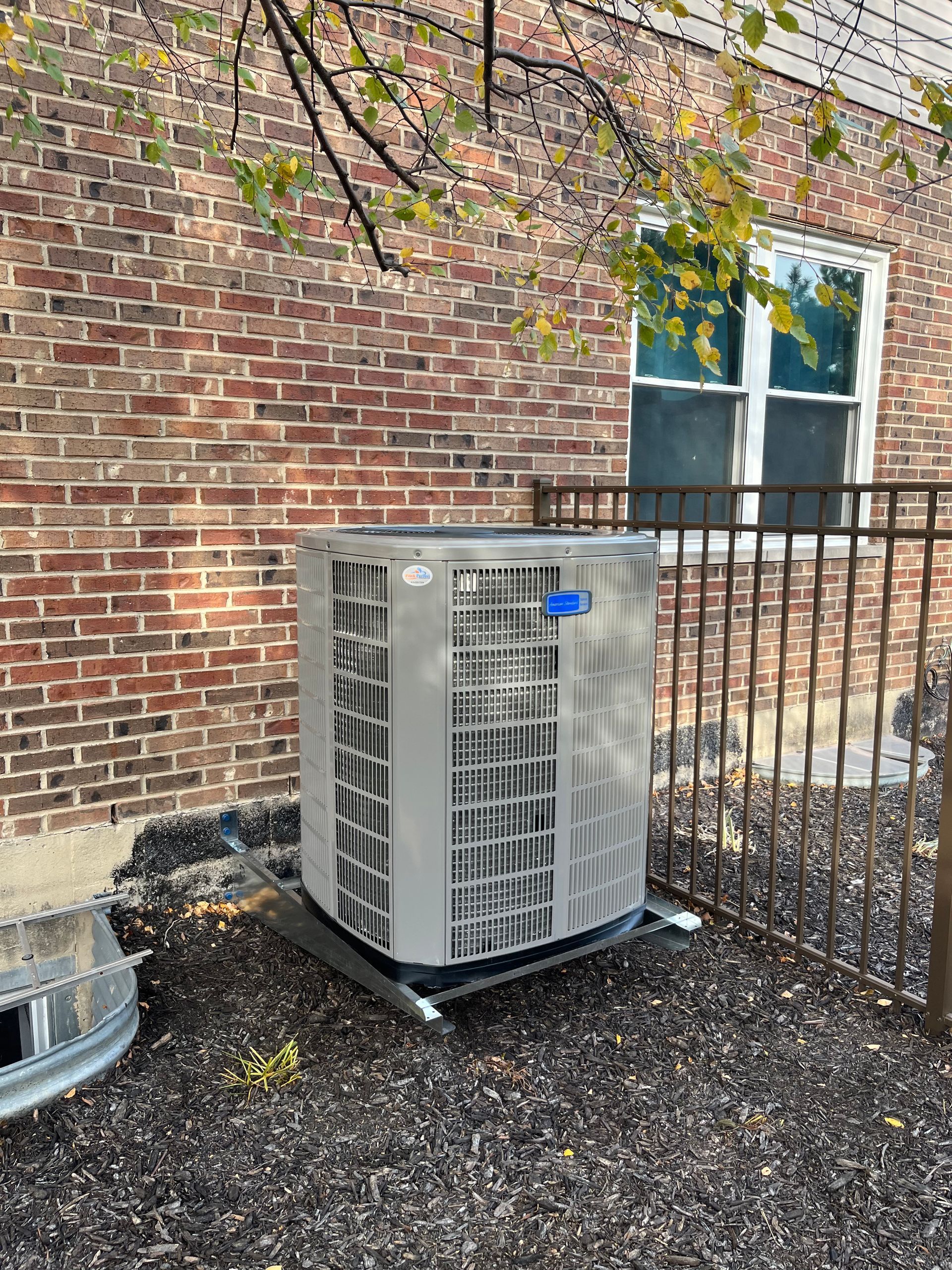 An air conditioning unit next to a brick building and a window, outdoors.