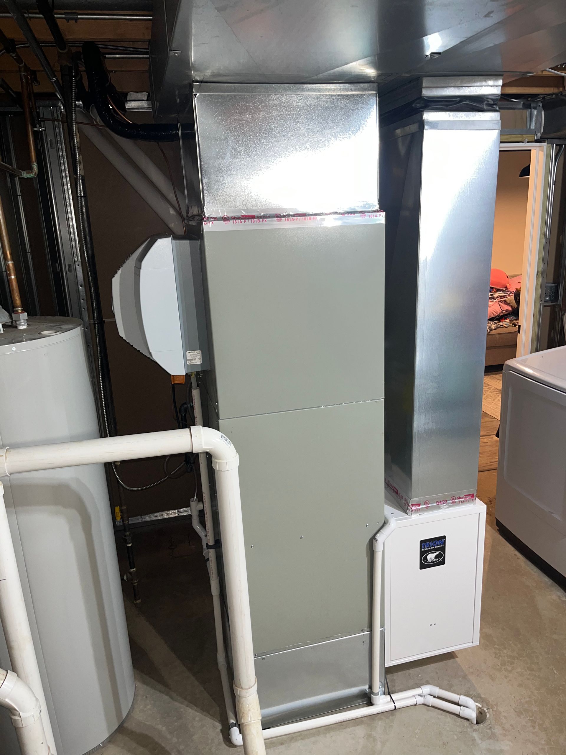 A stainless steel air conditioner is sitting in a basement next to a water heater.