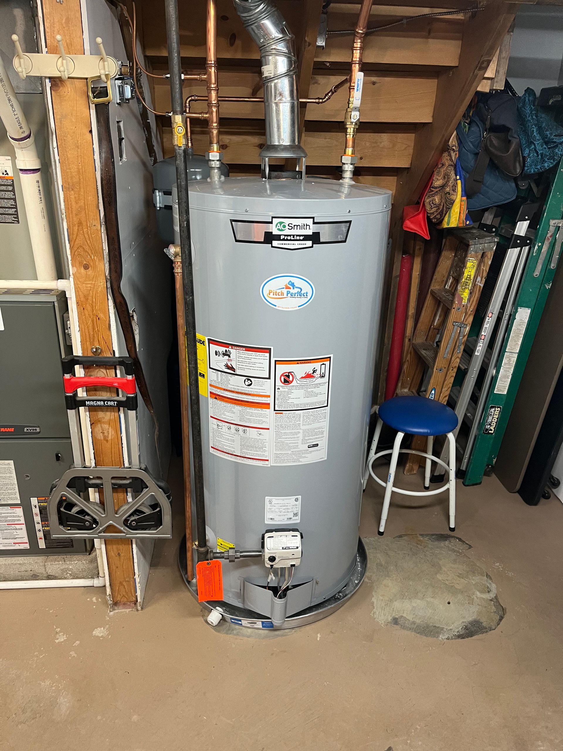 A gas water heater in a basement setting. It is grey, with copper piping and safety labels.