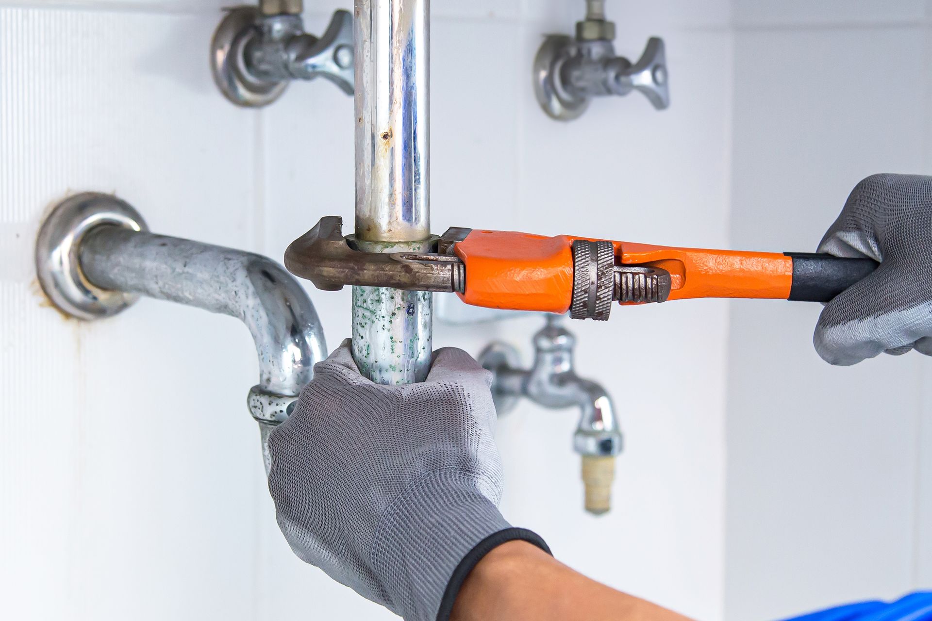 A plumber wearing gloves using an orange wrench to fix a pipe under a sink.