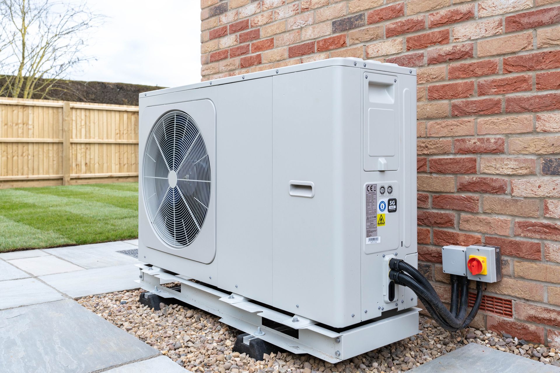 Heat pump unit outside brick wall, gray with black tubing, on gravel.