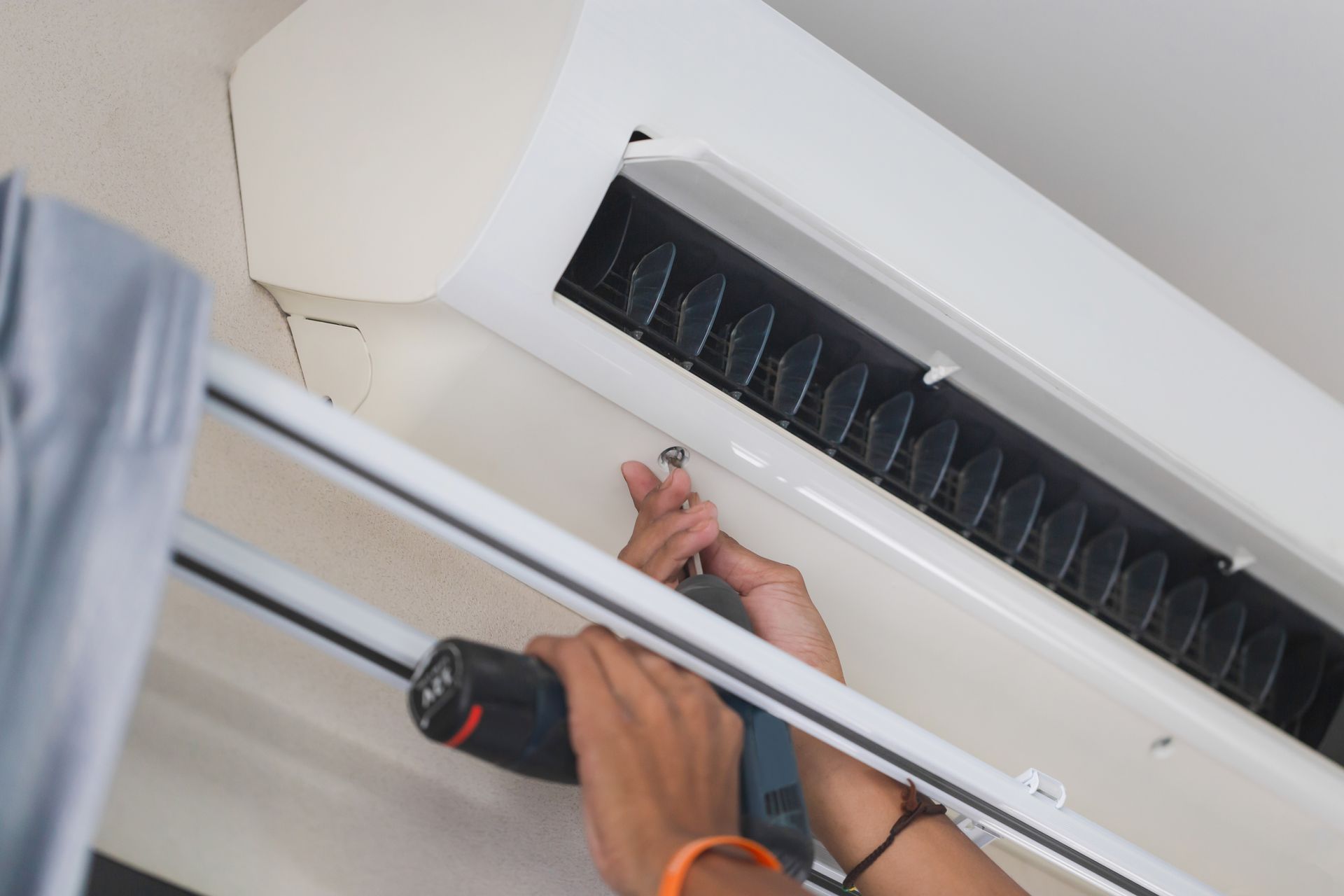 Person uses a power drill to work on a white air conditioning unit mounted to the ceiling.