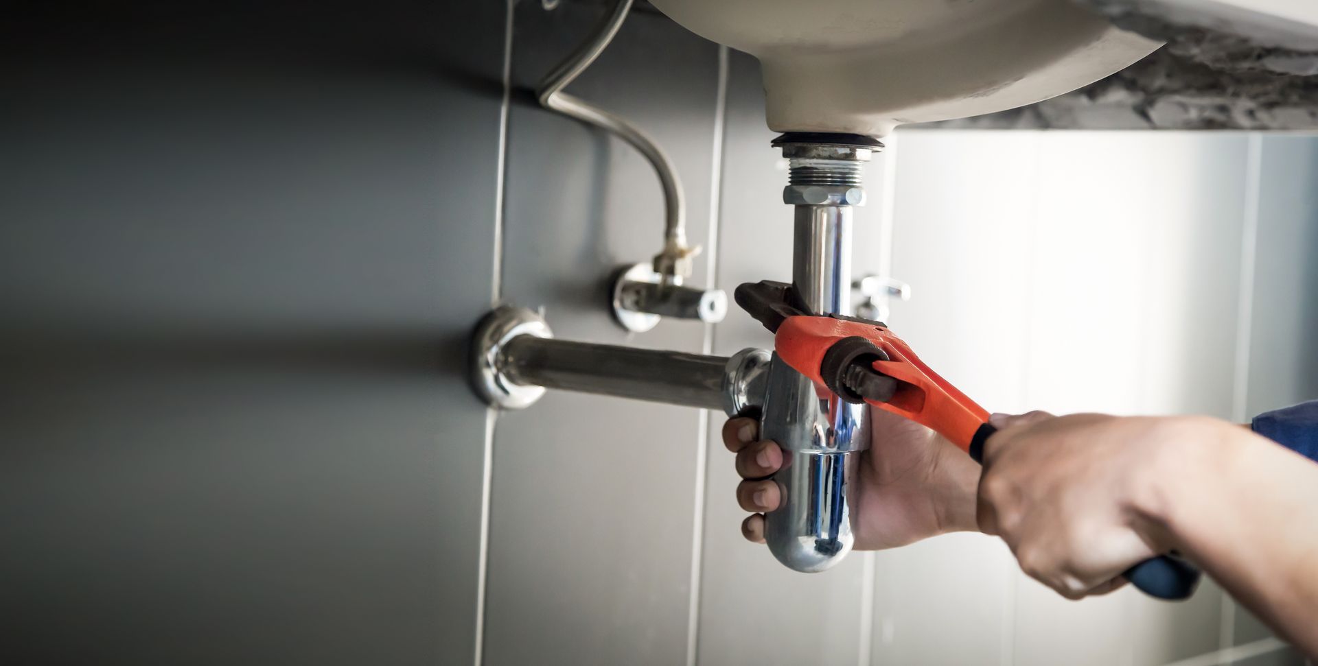 Plumber using a wrench to tighten a pipe under a bathroom sink against a gray tiled wall.