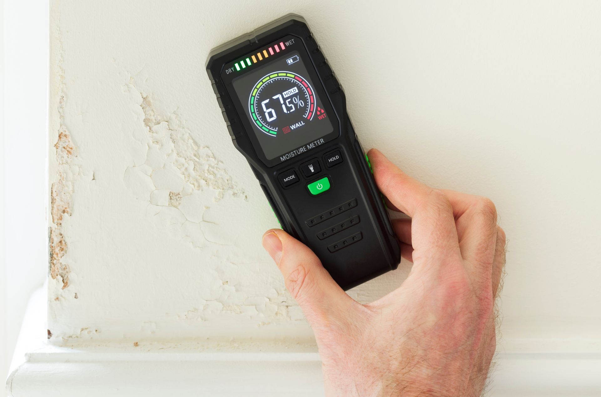 Hand holding a moisture meter against a damaged white wall, displaying a 67% moisture reading.
