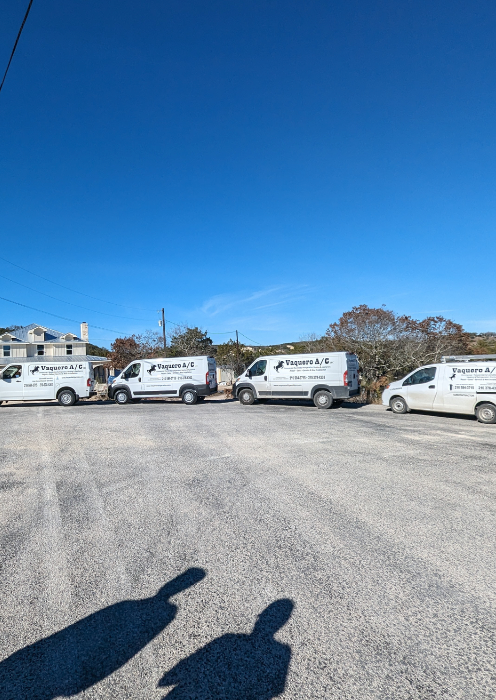 Four white vans parked in a gravel lot on a sunny day. Shadows of people are cast in foreground.