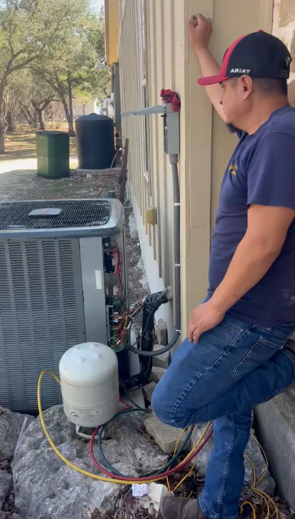 Man in cap leans on building, observing HVAC unit with connected hoses outdoors.