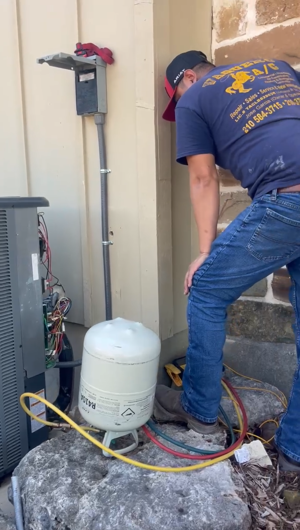 HVAC technician works on AC unit outside a building, with a tank and electrical box visible.