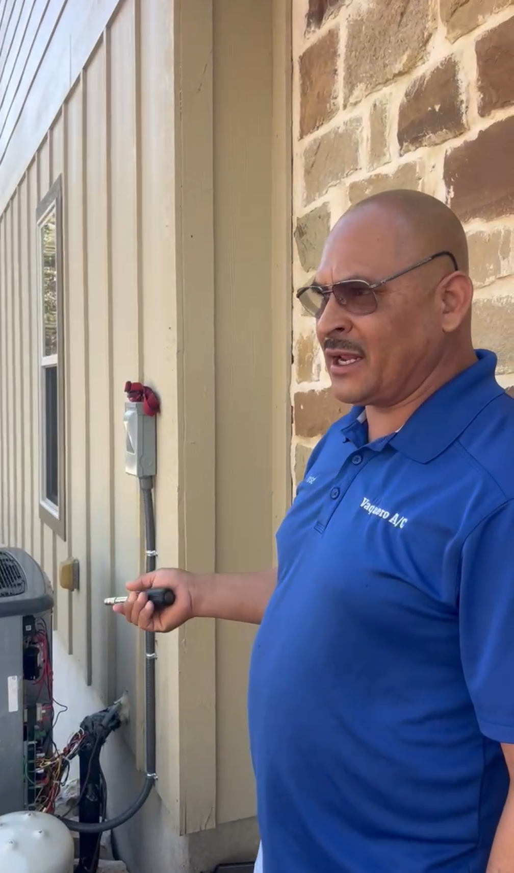 Man in blue shirt points to AC unit, outdoors. Building in background.