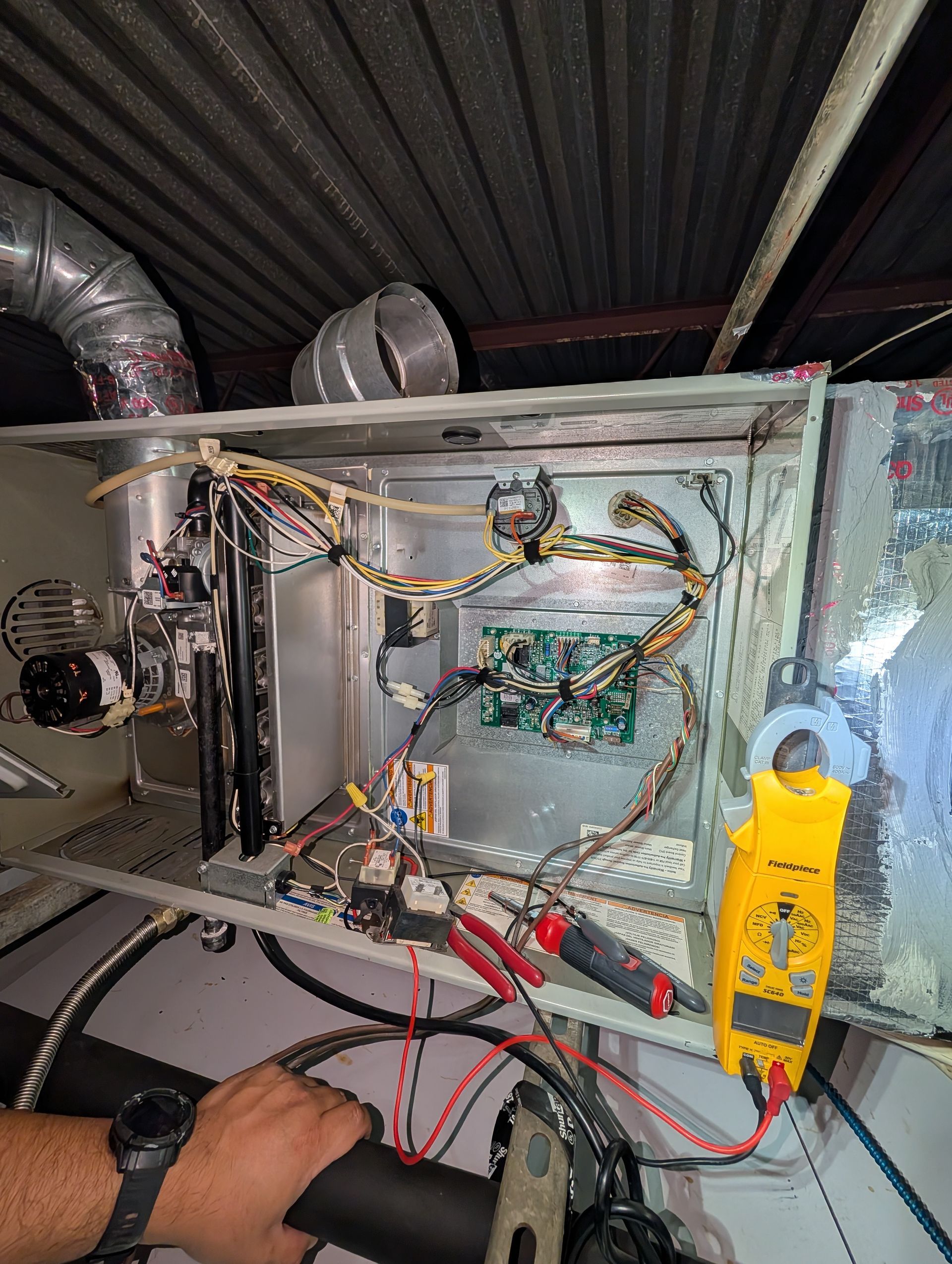 A technician testing an open HVAC furnace control panel with a yellow multimeter clamp meter in an attic.