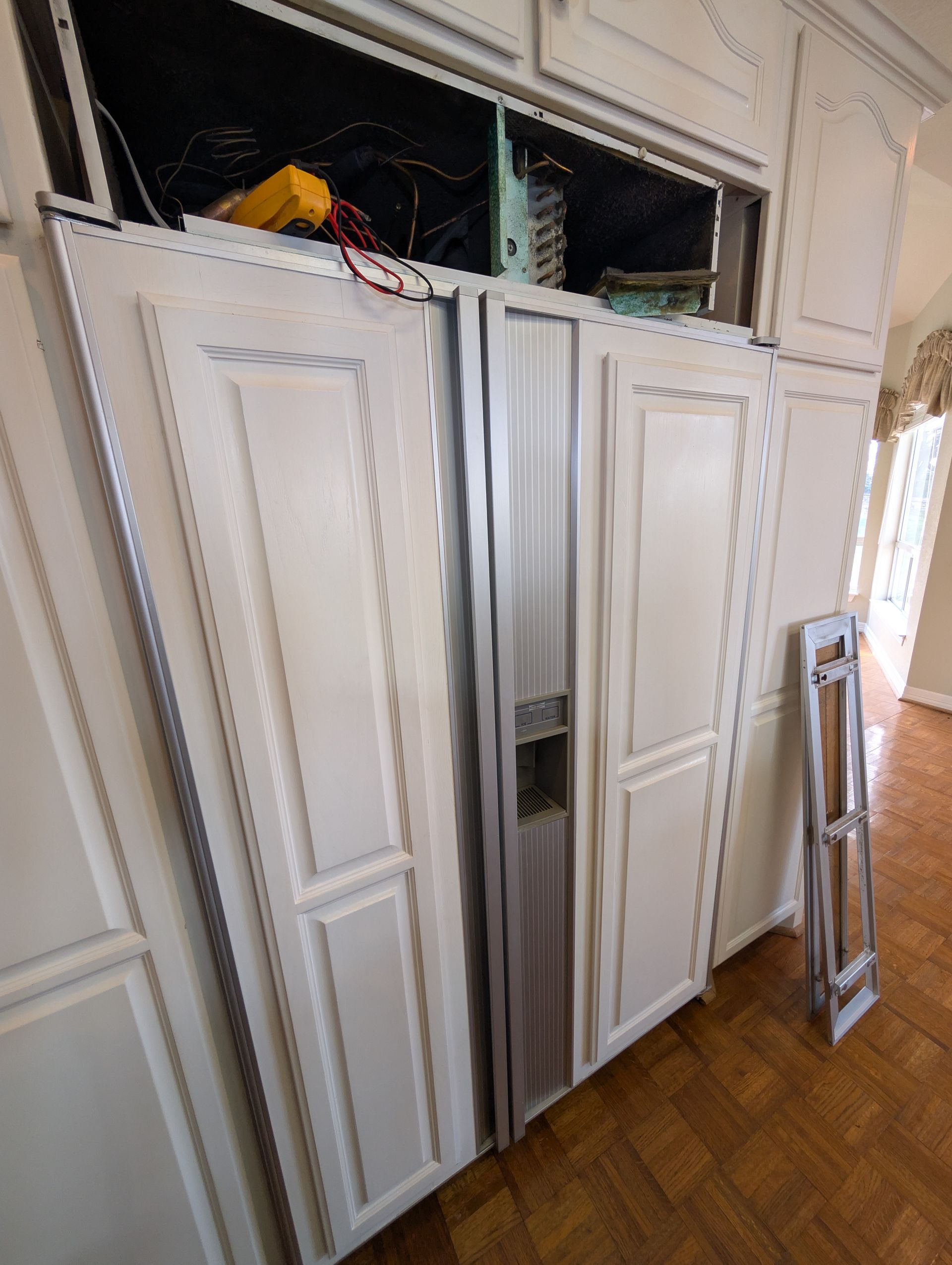 A built-in white refrigerator with its top panel removed, exposing wiring and a yellow tool inside a home kitchen.