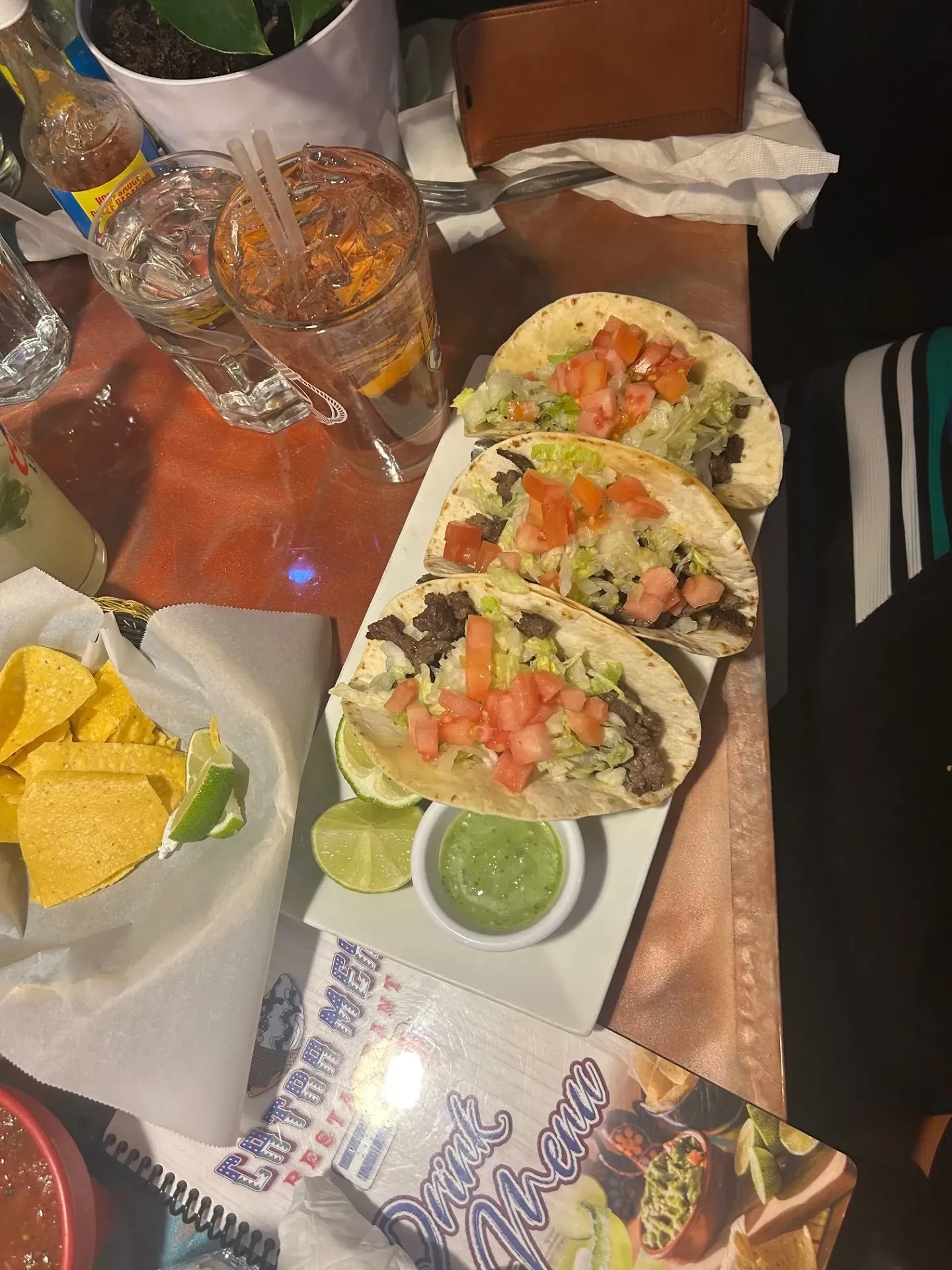 A table topped with tacos , chips , drinks and a menu.