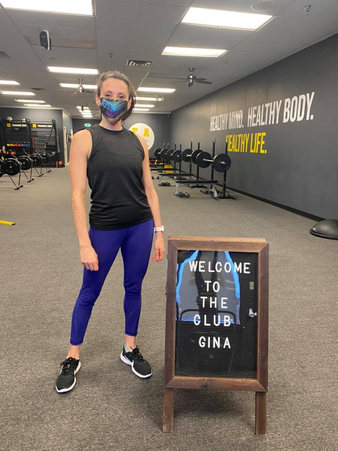 a woman wearing a mask is standing in a gym next to a sign that says welcome to the club gina .