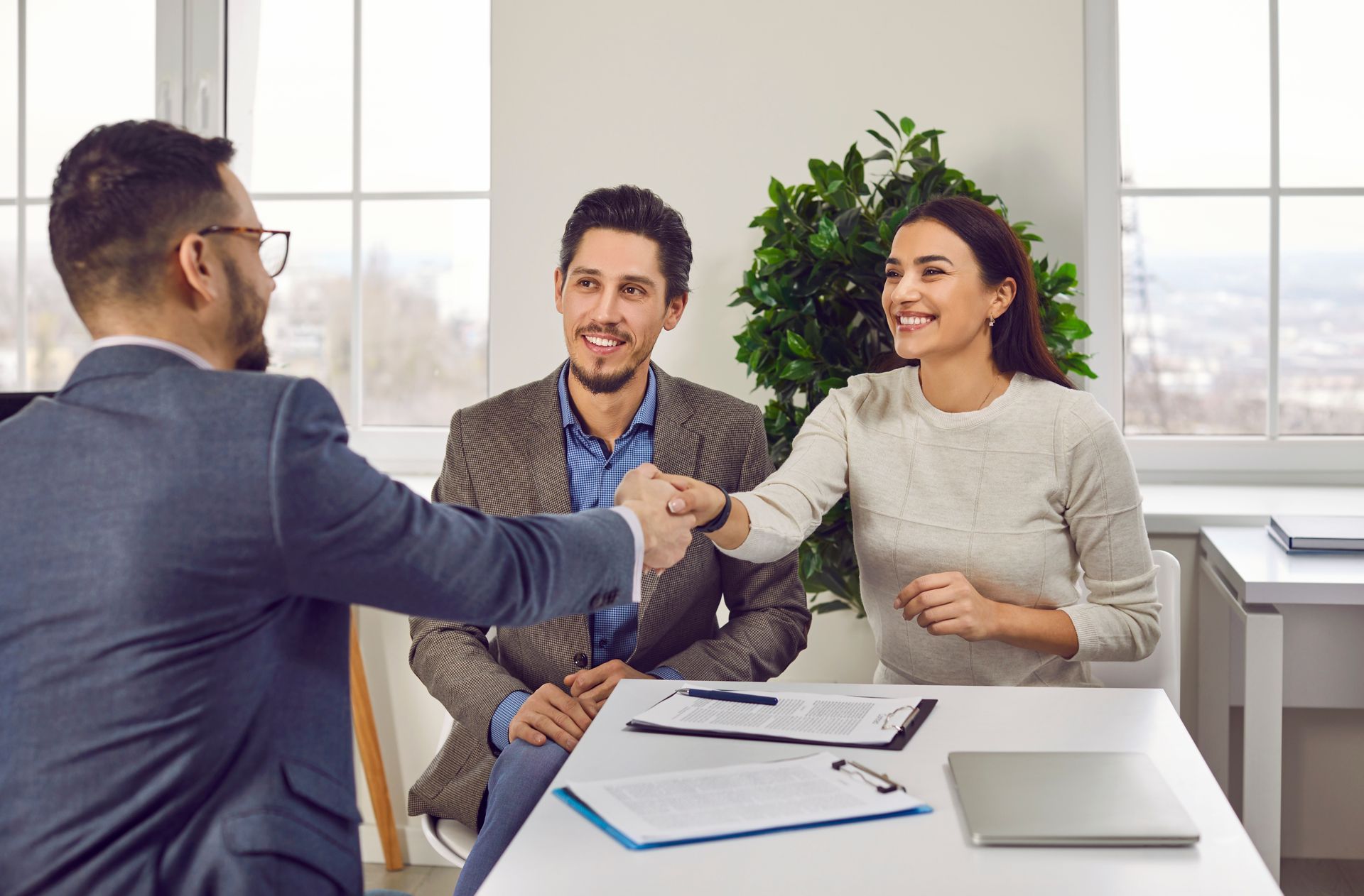 Three people in a bright office shake hands during a meeting, with papers and a laptop on the table.