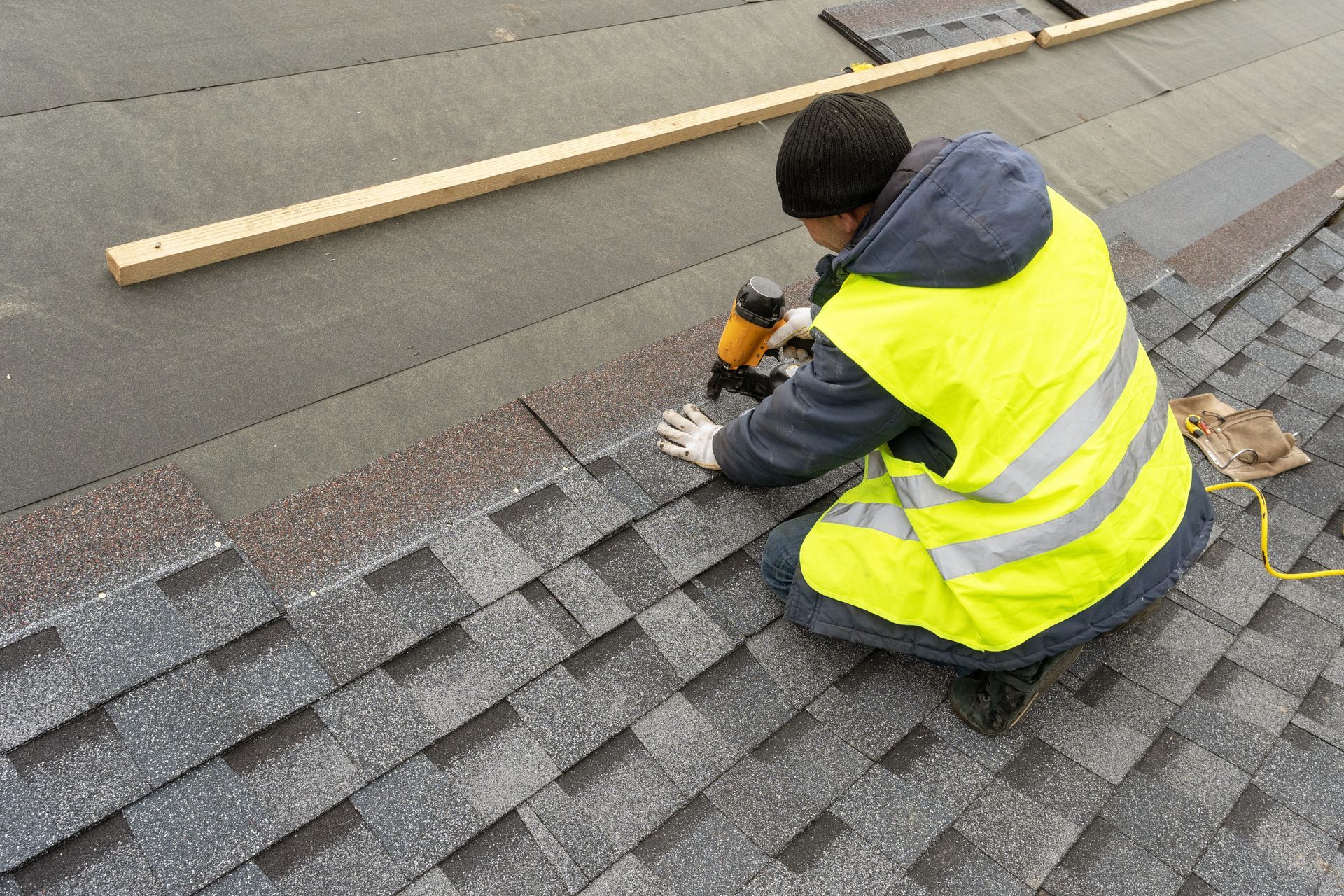 A roofer wearing a reflective jacket, performing a roofing install, and using a pneumatic nail gun.