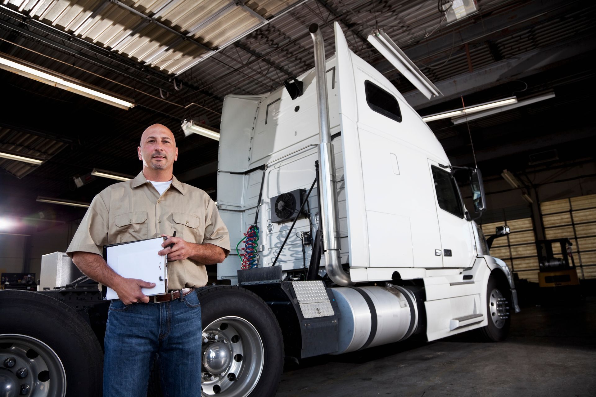 Truck driver standing next to semi-truck inside garage for heavy equipment transportation service. Truck driver standing next to semi-truck inside garage for heavy equipment transportation service.