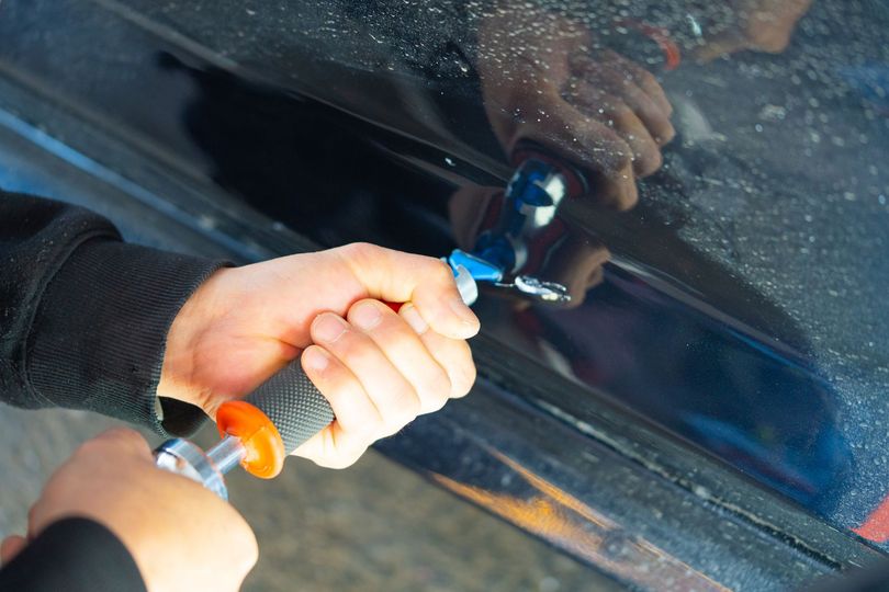 A hand using a suction-dent puller tool to remove a dent from a dark car door panel A hand using a suction-dent puller tool to remove a dent from a dark car door panel
