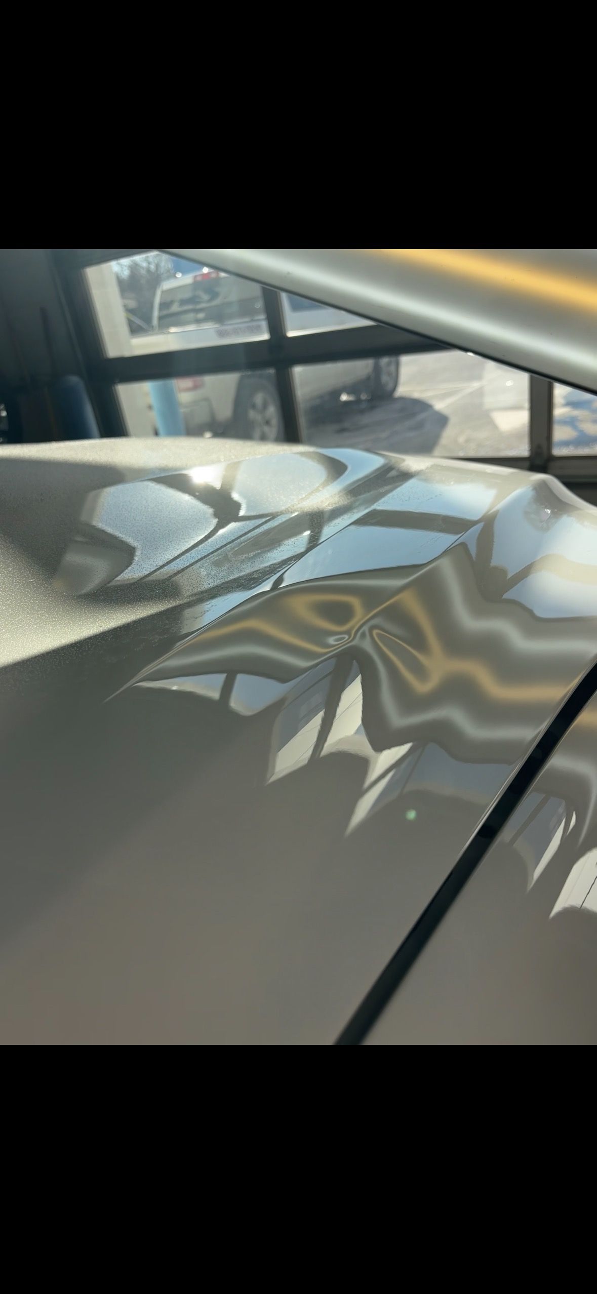 Close-up of a damaged, light-colored car hood with a large dent reflecting the sky and windows.