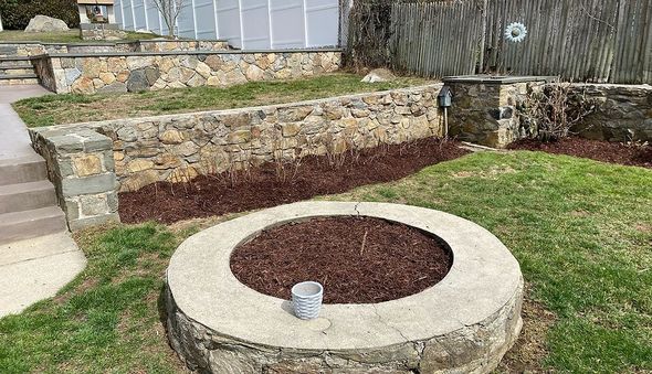 A round stone-walled garden bed with dark mulch and a small planter sits in a backyard near tiered stone retaining walls. A round stone-walled garden bed with dark mulch and a small planter sits in a backyard near tiered stone retaining walls.