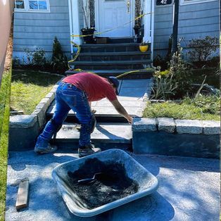 A person in a red shirt and blue jeans works on stone steps in front of a house, with a wheelbarrow in the foreground.
