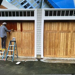 A person standing on a ladder stains the left wooden garage door a dark brown color, while the right door remains natural.