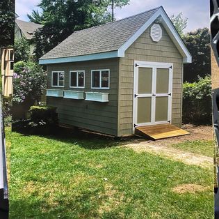 A tan storage shed with white trim, three windows with flower boxes, and a ramp leading to double doors in a grassy yard.