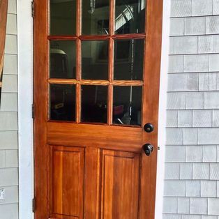 A wooden exterior door with nine glass window panes and two lower panels, installed in a home with grey shingle siding.