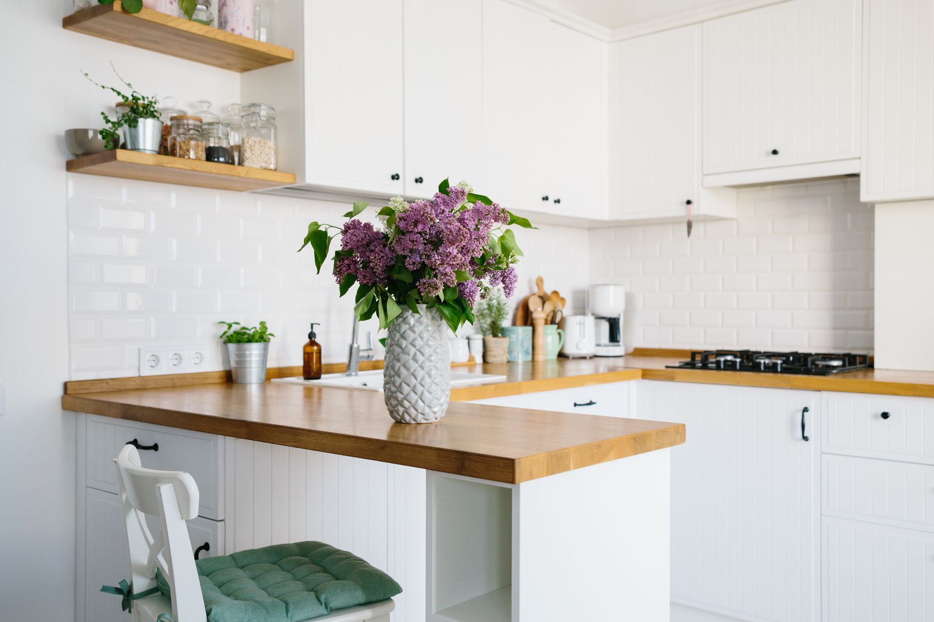 A clean white kitchen with wooden countertops, a vase of purple lilacs, floating shelves, and a white chair with a cushion. A clean white kitchen with wooden countertops, a vase of purple lilacs, floating shelves, and a white chair with a cushion.