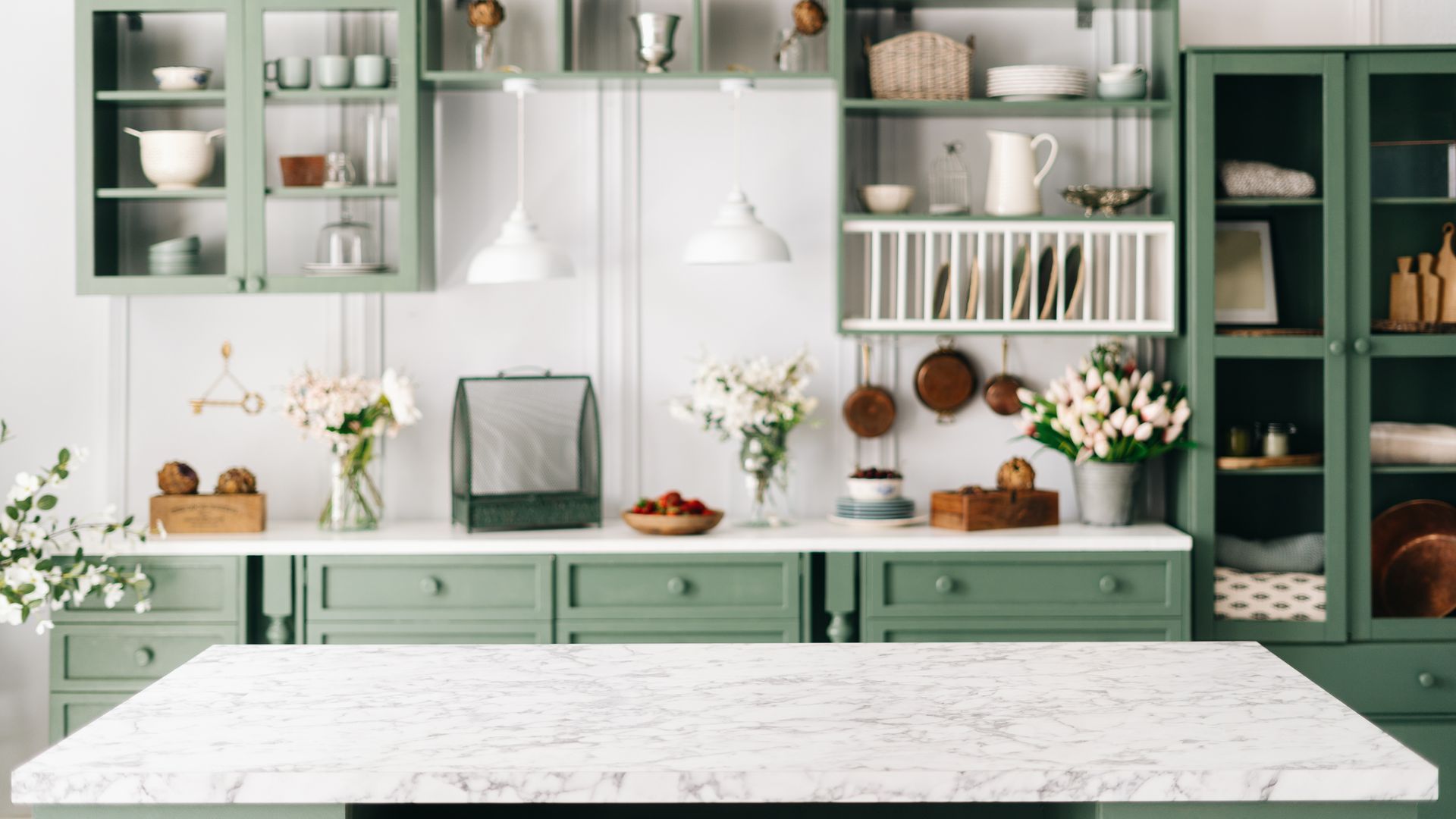 A bright, modern kitchen featuring sage green cabinets, a white marble countertop, open shelving, and vases of flowers. A bright, modern kitchen featuring sage green cabinets, a white marble countertop, open shelving, and vases of flowers.