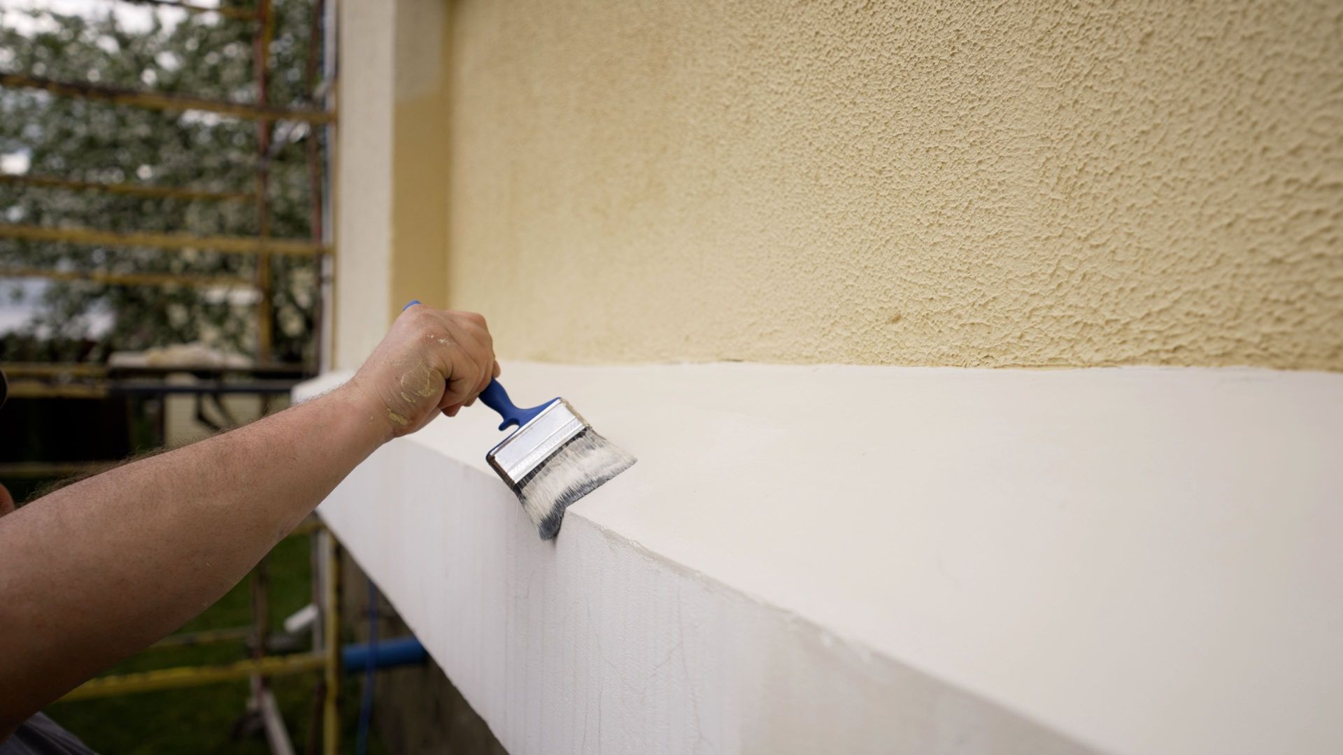 A person uses a brush to paint a white horizontal ledge against a textured yellow wall outdoors.