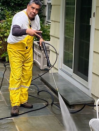 A person in yellow rain overalls uses a pressure washer to clean a stone patio next to a house. A person in yellow rain overalls uses a pressure washer to clean a stone patio next to a house.