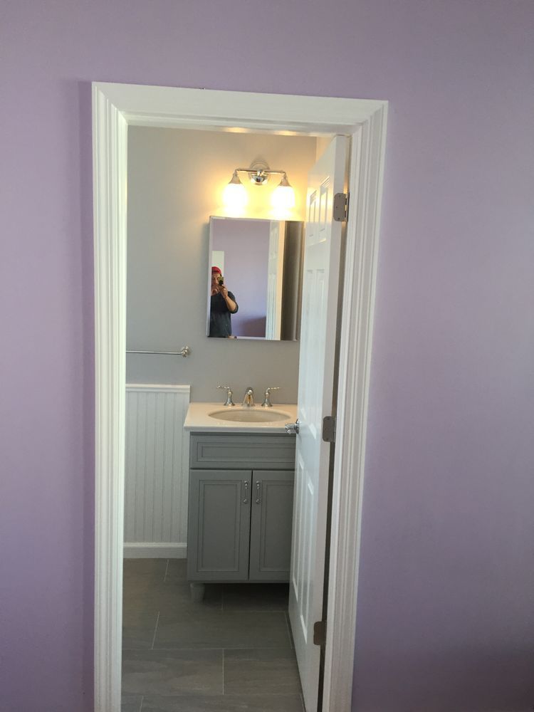 A view through a white door frame into a small bathroom with gray walls, a vanity, and an illuminated mirror. A view through a white door frame into a small bathroom with gray walls, a vanity, and an illuminated mirror.