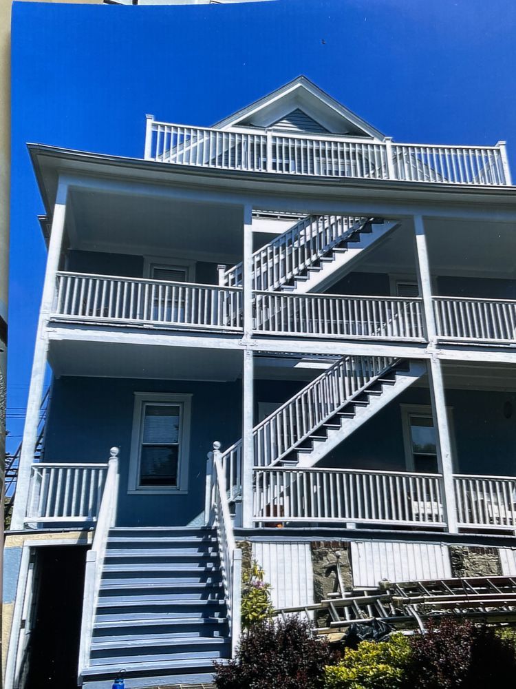 A three-story blue house with white railings, featuring external staircases connecting the levels under a clear blue sky.