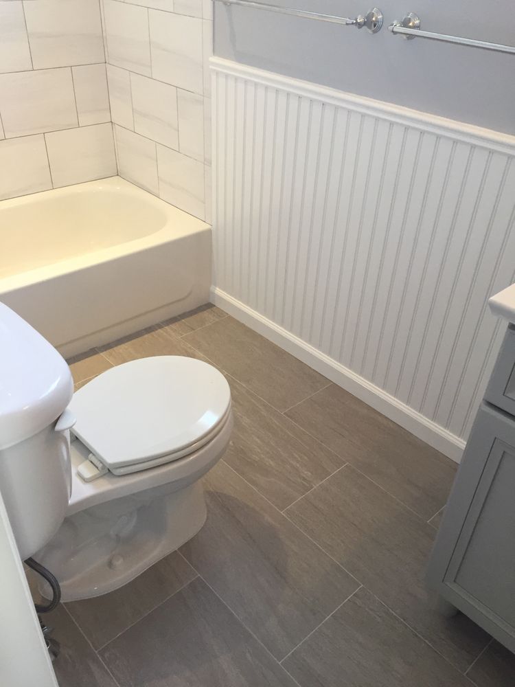 A modern bathroom with a white toilet, gray wood-look tile flooring, white beadboard wainscoting, and a white bathtub. A modern bathroom with a white toilet, gray wood-look tile flooring, white beadboard wainscoting, and a white bathtub.