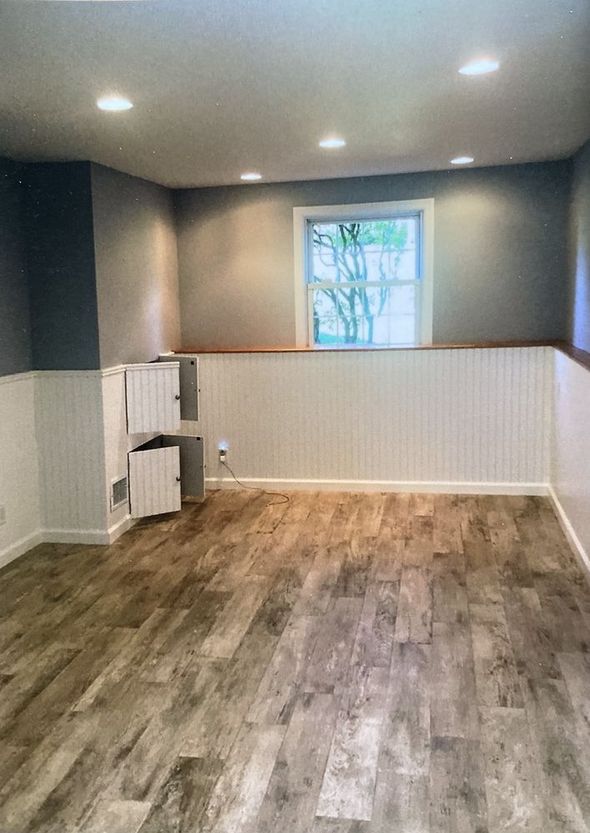 A room with gray walls, white wainscoting, a centered window, and wood-look flooring with a small white cabinet. A room with gray walls, white wainscoting, a centered window, and wood-look flooring with a small white cabinet.