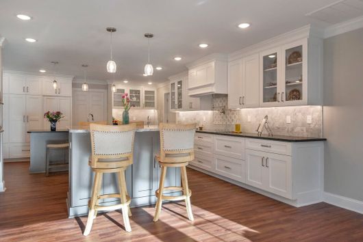 A kitchen with white cabinets, wooden floors, stools, and a large island.