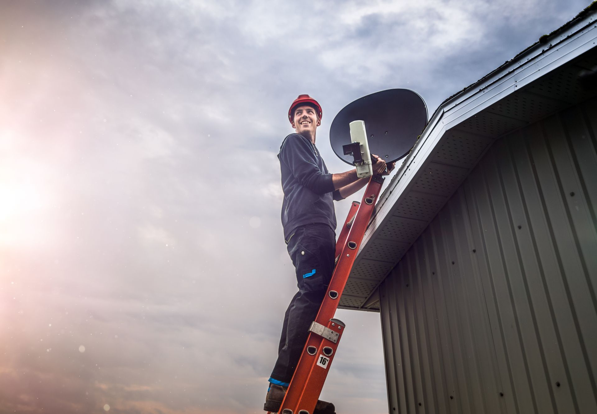 A man is standing on a ladder installing a satellite dish on the roof of a building.
