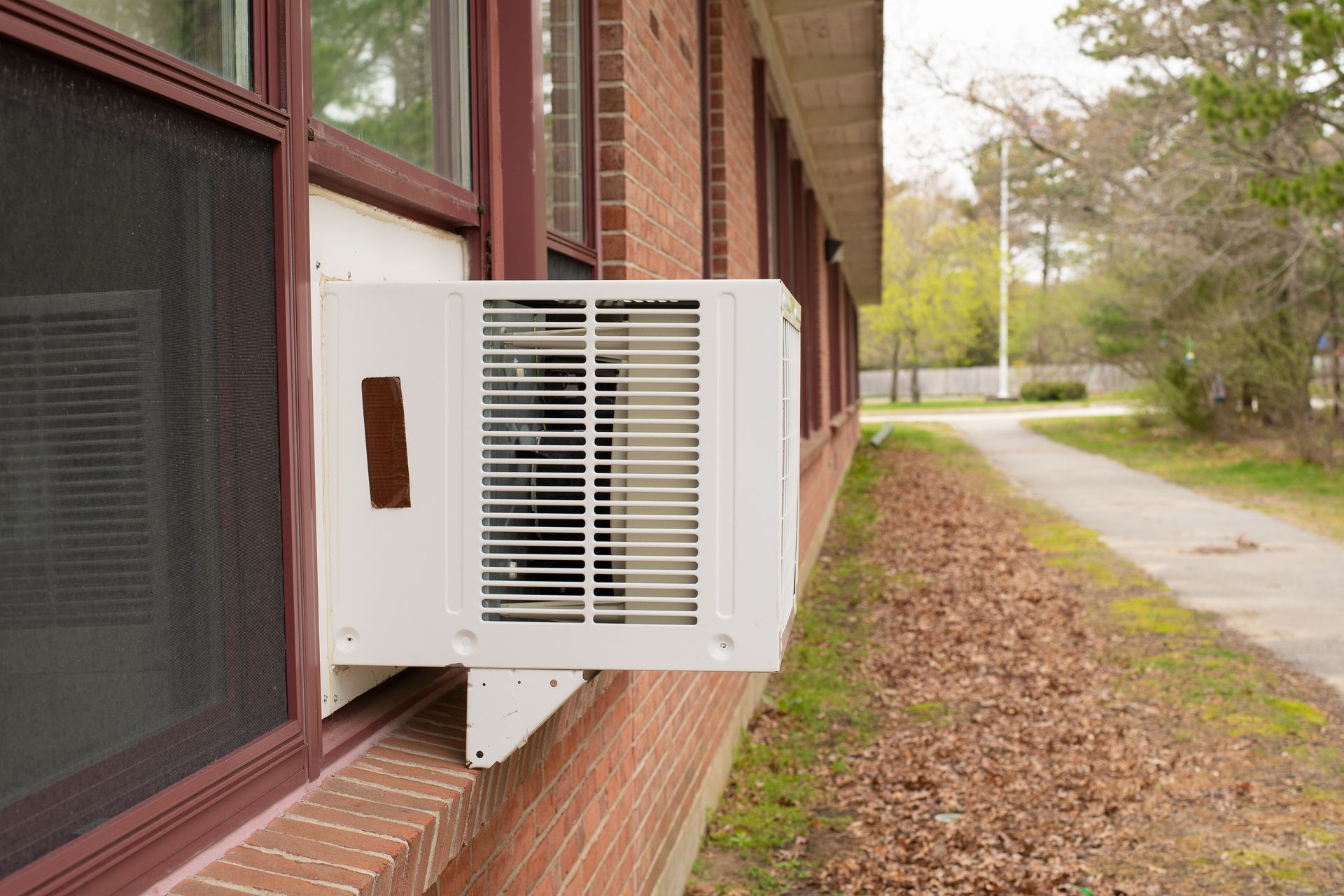 White air conditioner in a brick building window, near a sidewalk and trees.