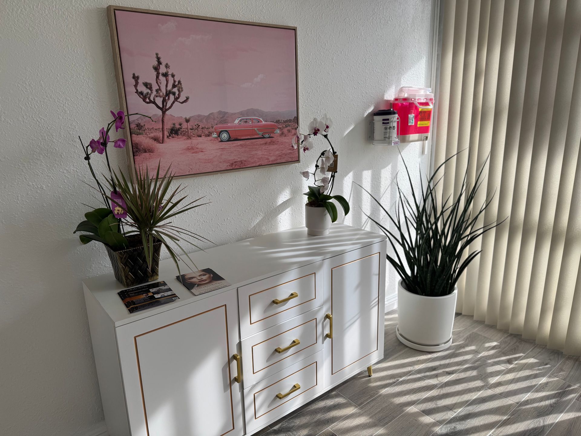 A room with a dresser , two potted plants and a painting on the wall.