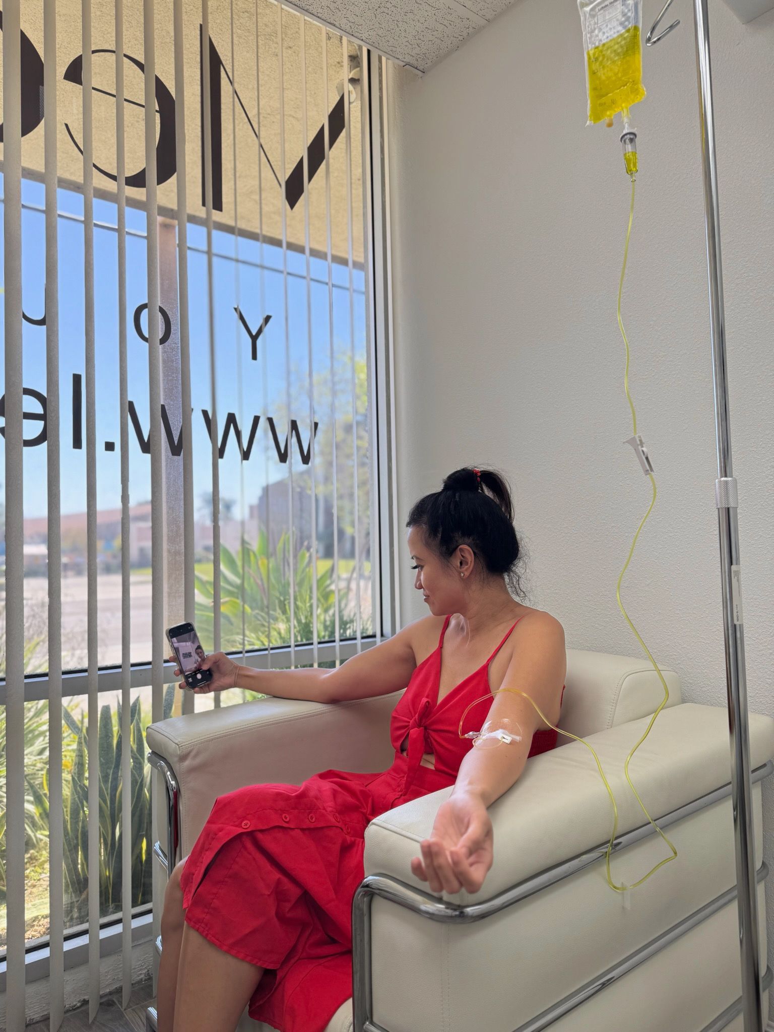 Woman in red dress receiving IV drip, seated by a window, holding phone.