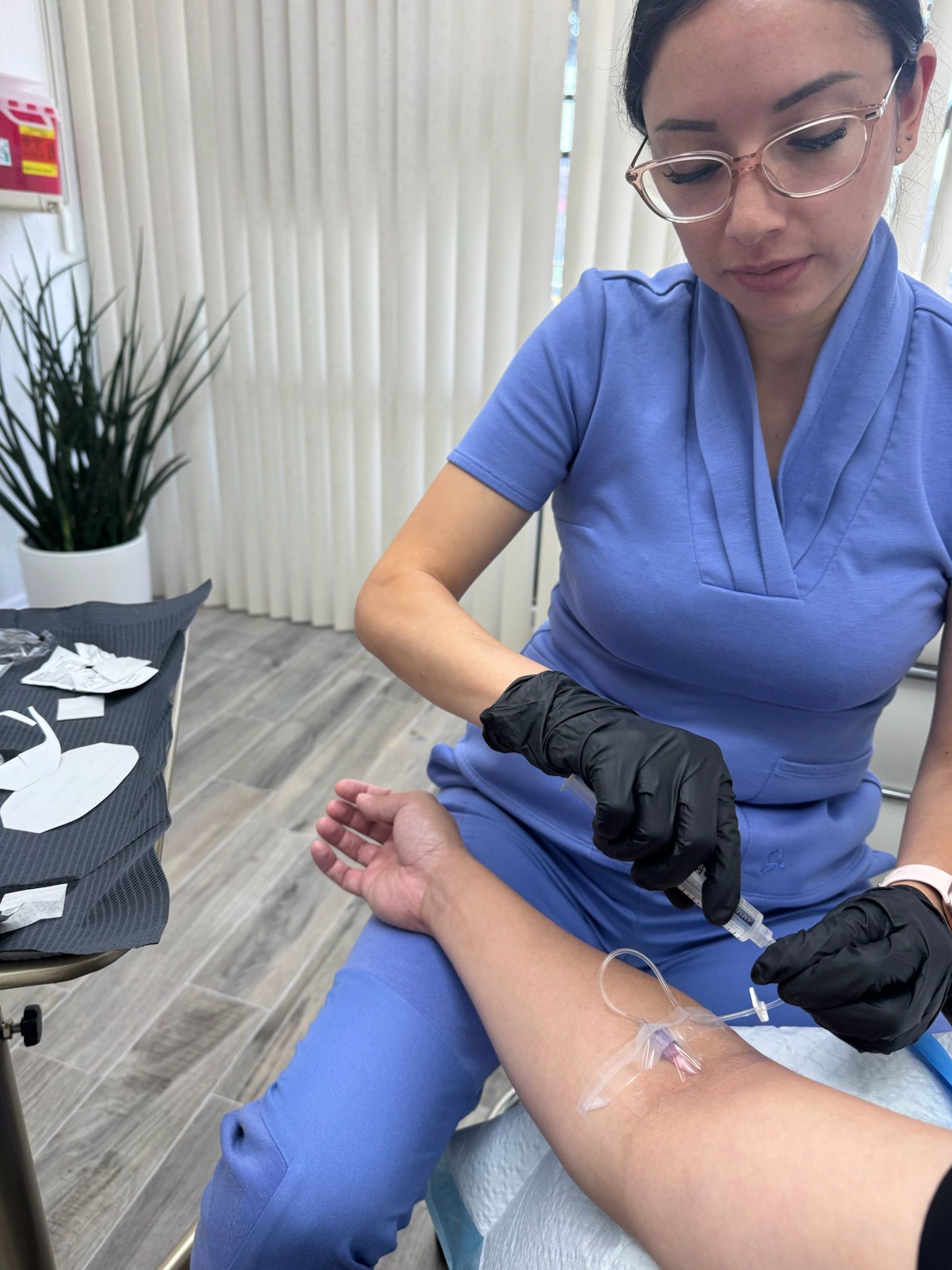 A medical professional prepares an IV on a patient's arm. She wears gloves and scrubs.