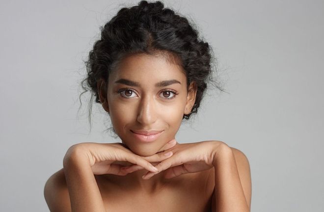 A young woman with curly hair is smiling with her hands on her face.