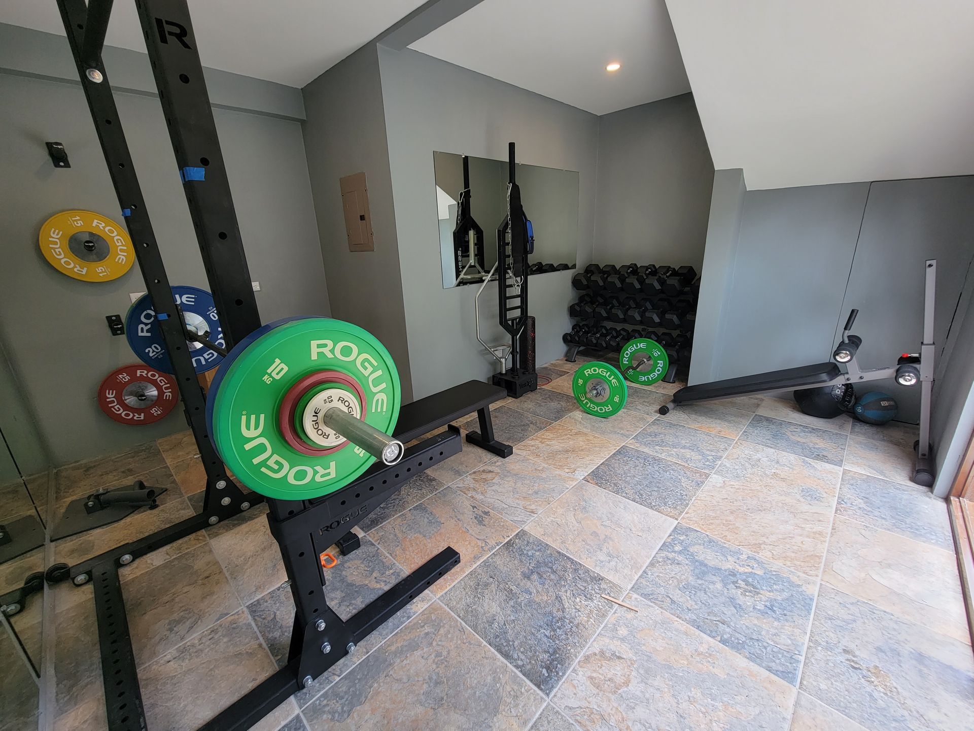 A gym with a barbell rack , bench , dumbbells , and a mirror.