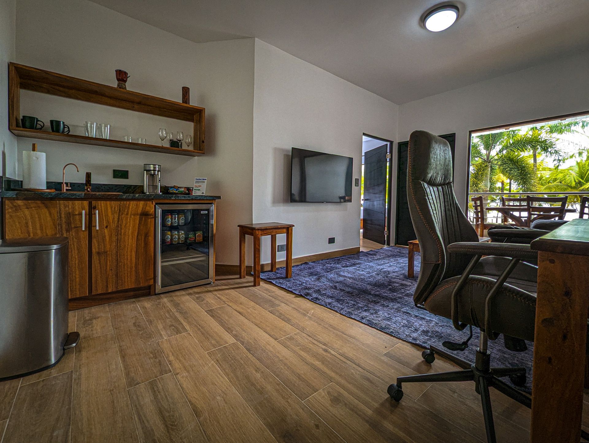 A living room with hardwood floors , a desk , chairs , a refrigerator and a flat screen tv.