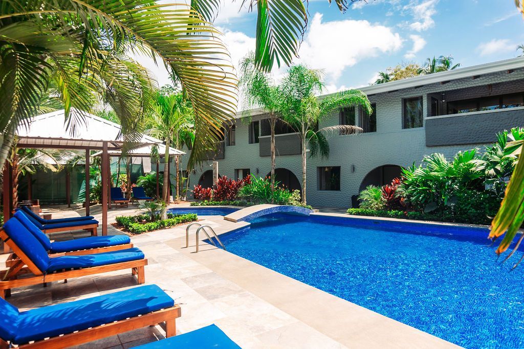A large swimming pool surrounded by chairs and palm trees in front of a building.